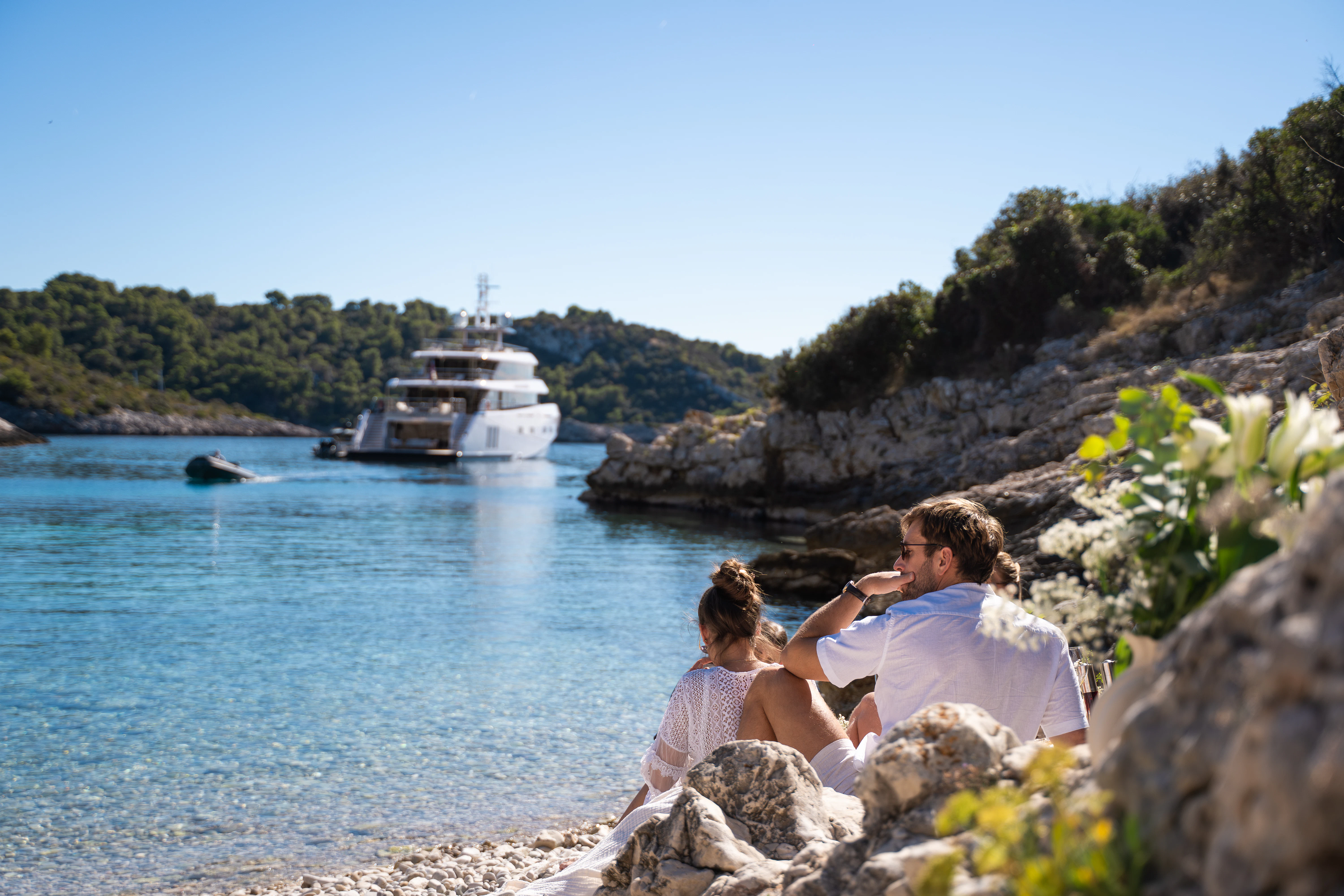 a man and woman sitting on a rock by the water with a boat in the background aboard RENATA Yacht for Charter