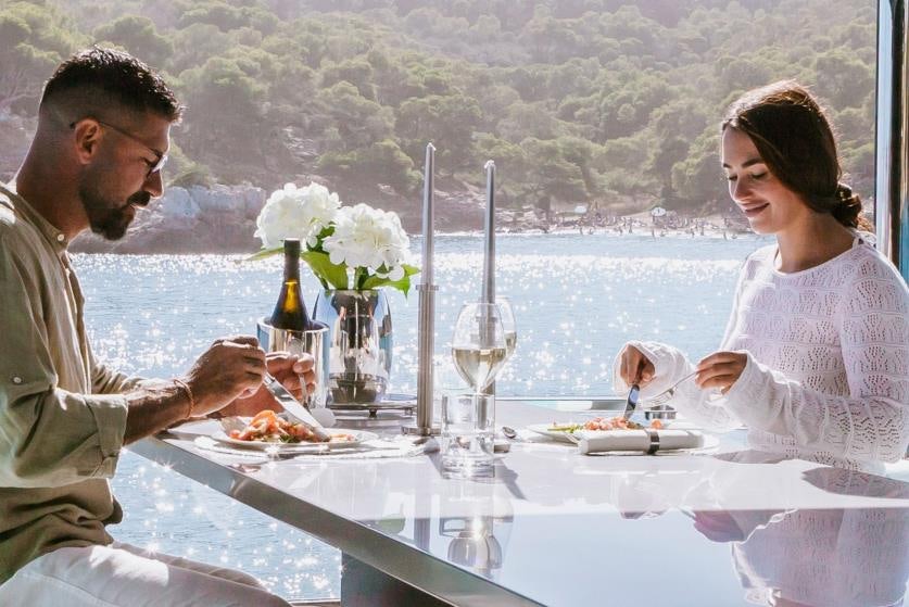 a man and a woman eating at a table with a lake and mountains in the background aboard BARRACUDA III Yacht for Charter