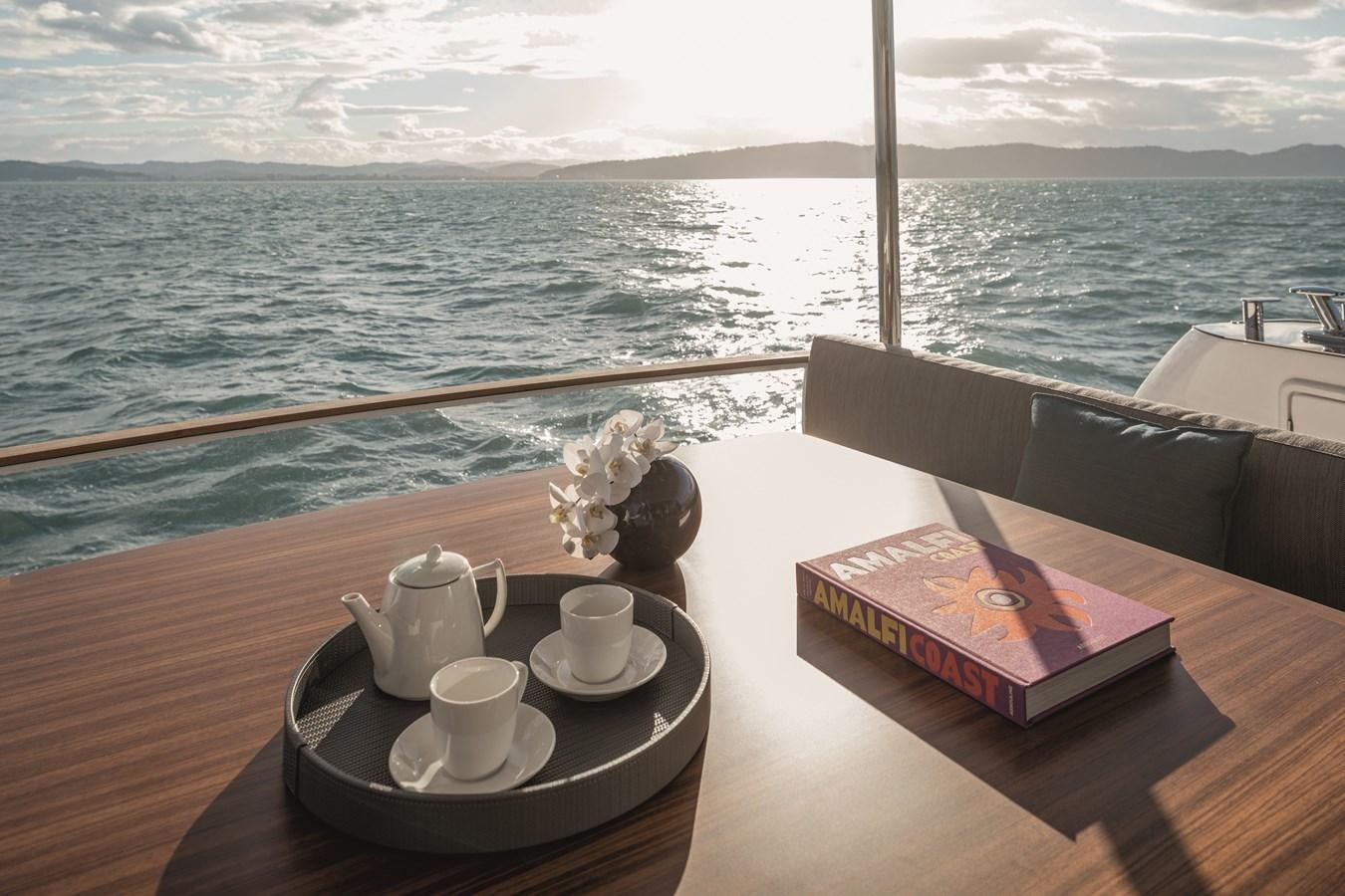 a table with cups and a book on it by a window with a view of the ocean and aboard 2023 MAGELLANO 66 Yacht for Sale