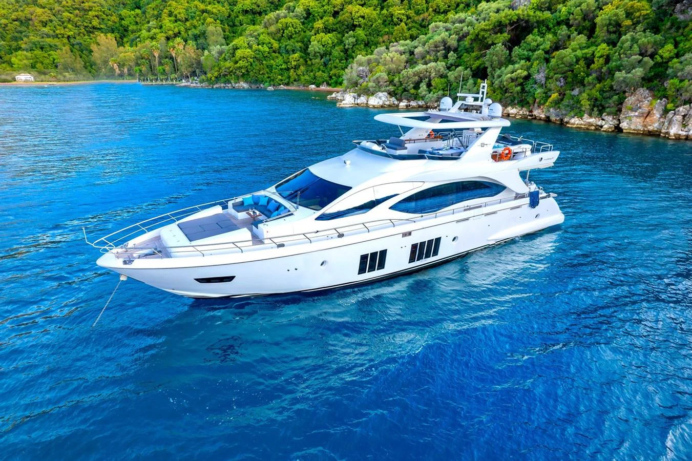 A sleek, modern yacht cruising in clear blue waters near a lush, green coastline. aboard HUBO Yacht for Sale