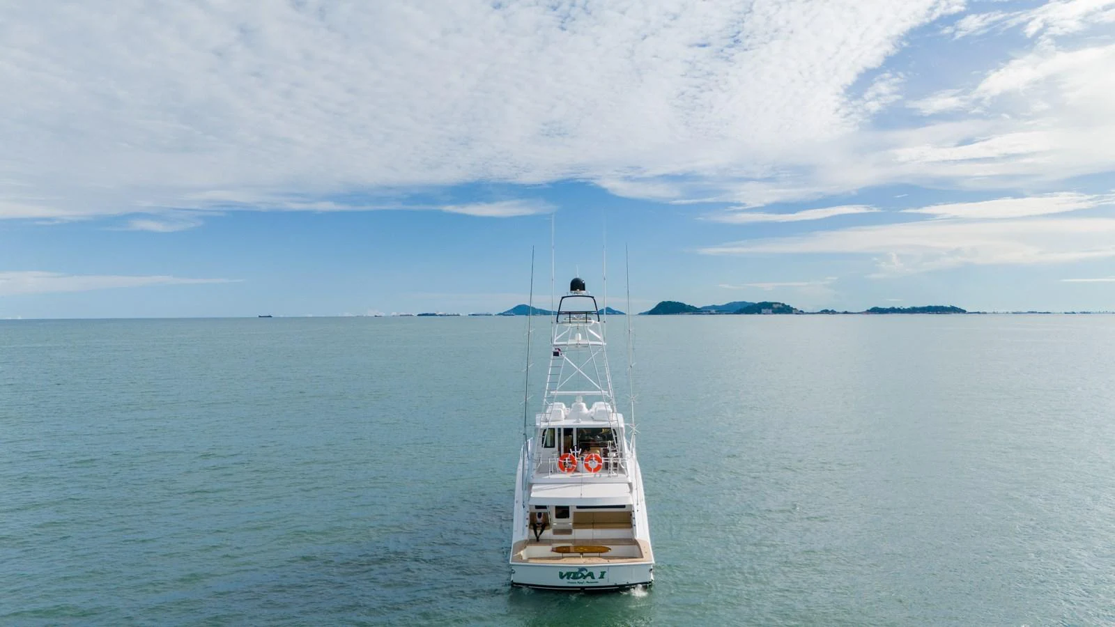 The yacht is seen from the rear, cruising on calm waters under a bright, partly cloudy sky. aboard VIDA 1 Yacht for Sale