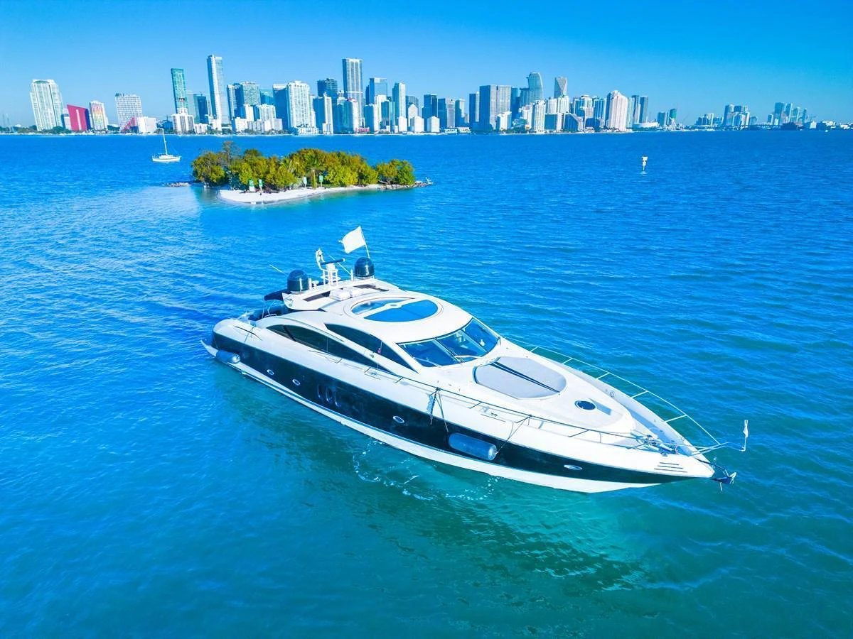 A sleek yacht glides through vibrant blue waters with a city skyline in the background. aboard THE GAMBLER Yacht for Sale