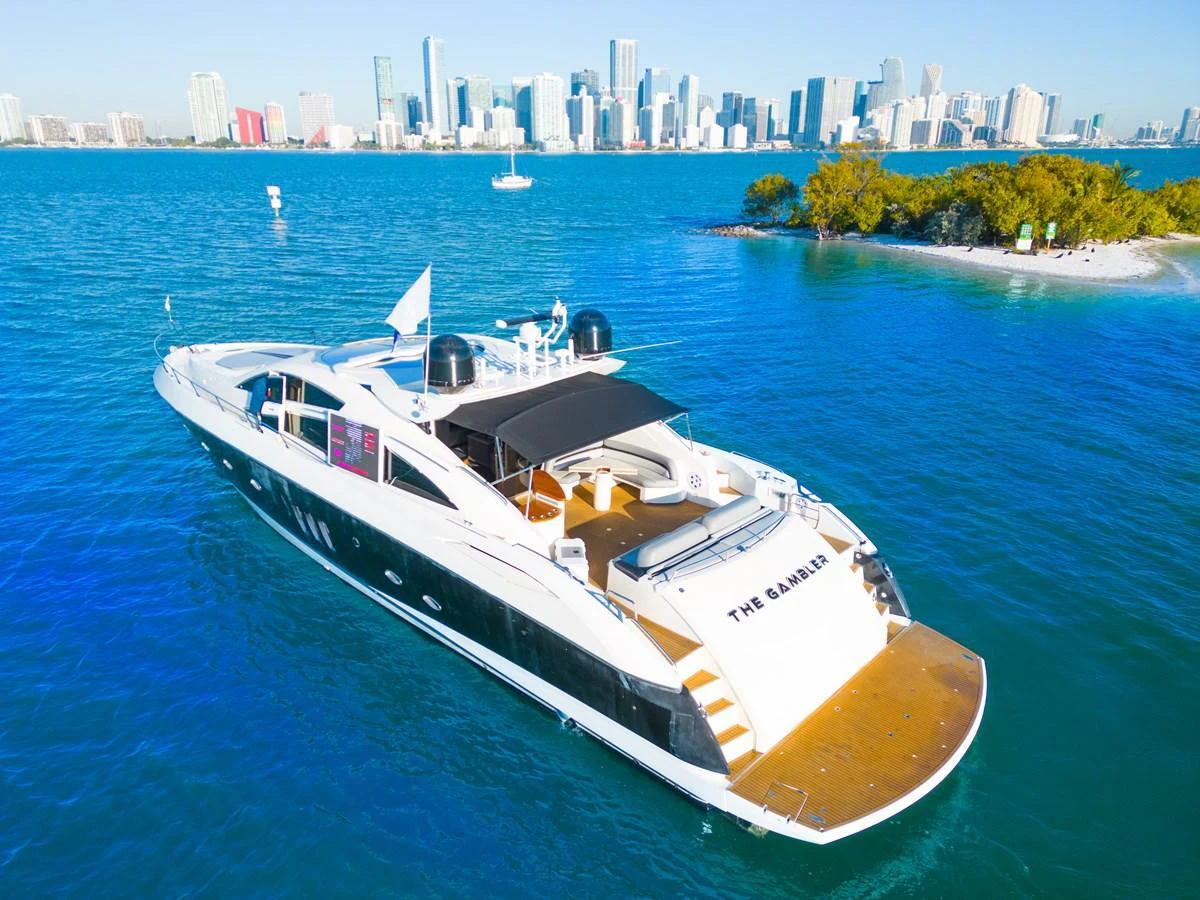 A sleek and modern yacht cruises through vibrant blue waters with a city skyline in the background. aboard THE GAMBLER Yacht for Sale