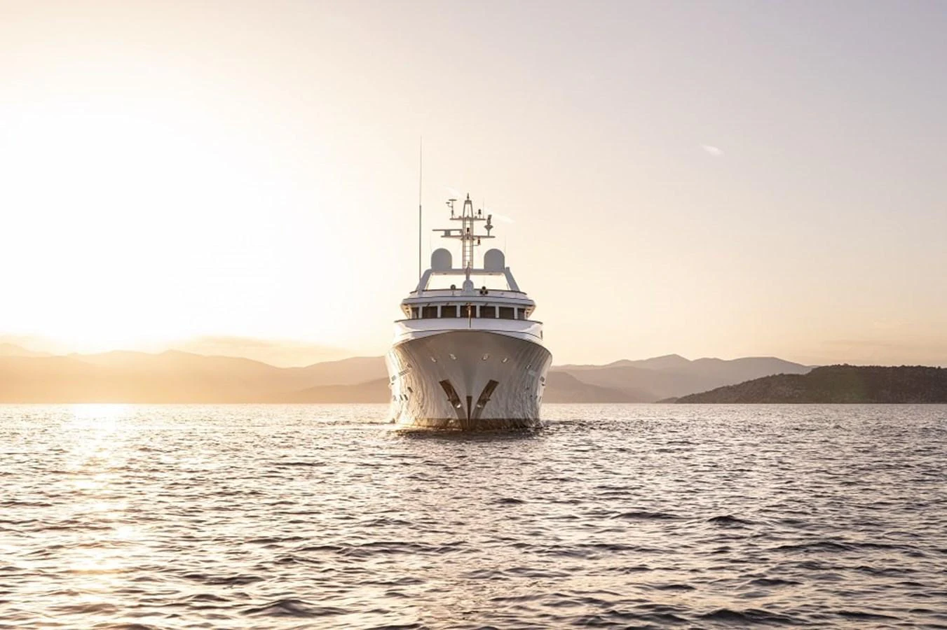 A majestic yacht sails towards the camera, bathed in the golden light of sunset over calm waters. aboard KASSANDRA Yacht for Sale