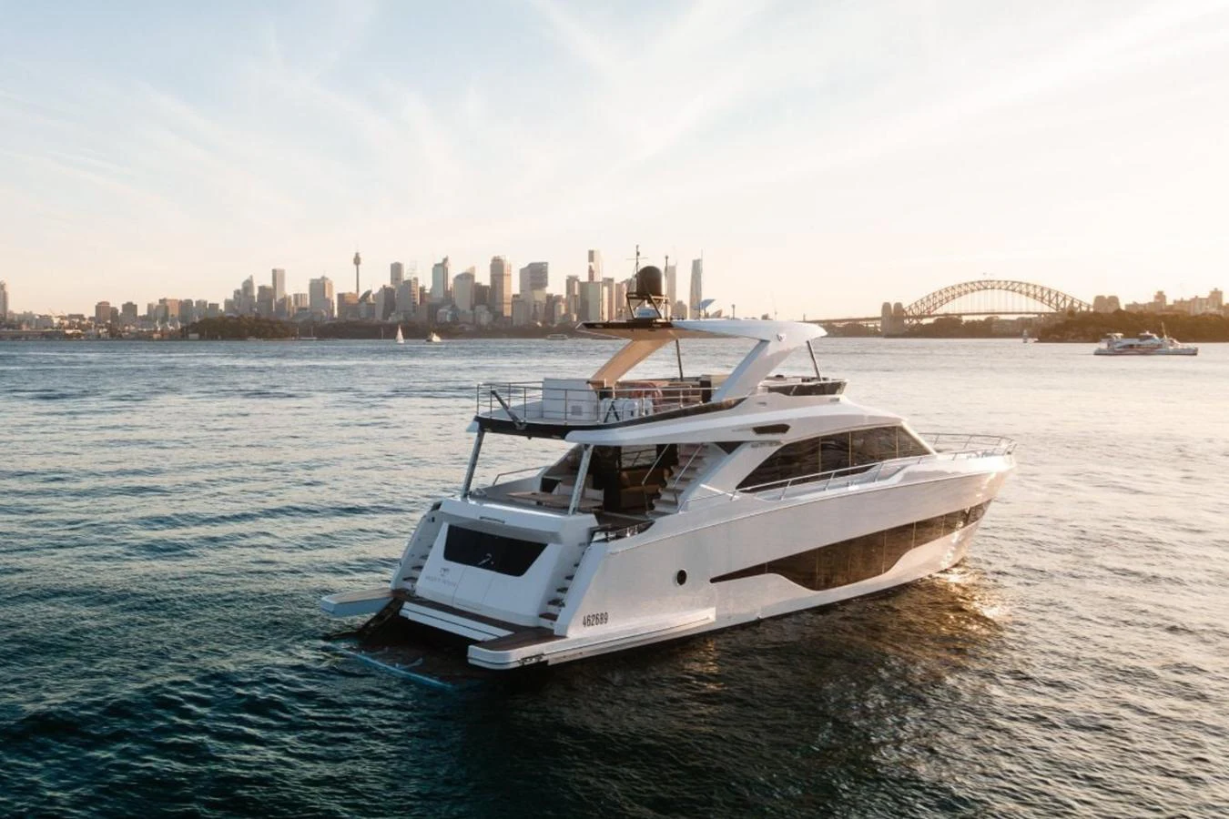 A sleek yacht cruises through calm waters with a cityscape in the background. aboard TOPAZ Yacht for Sale