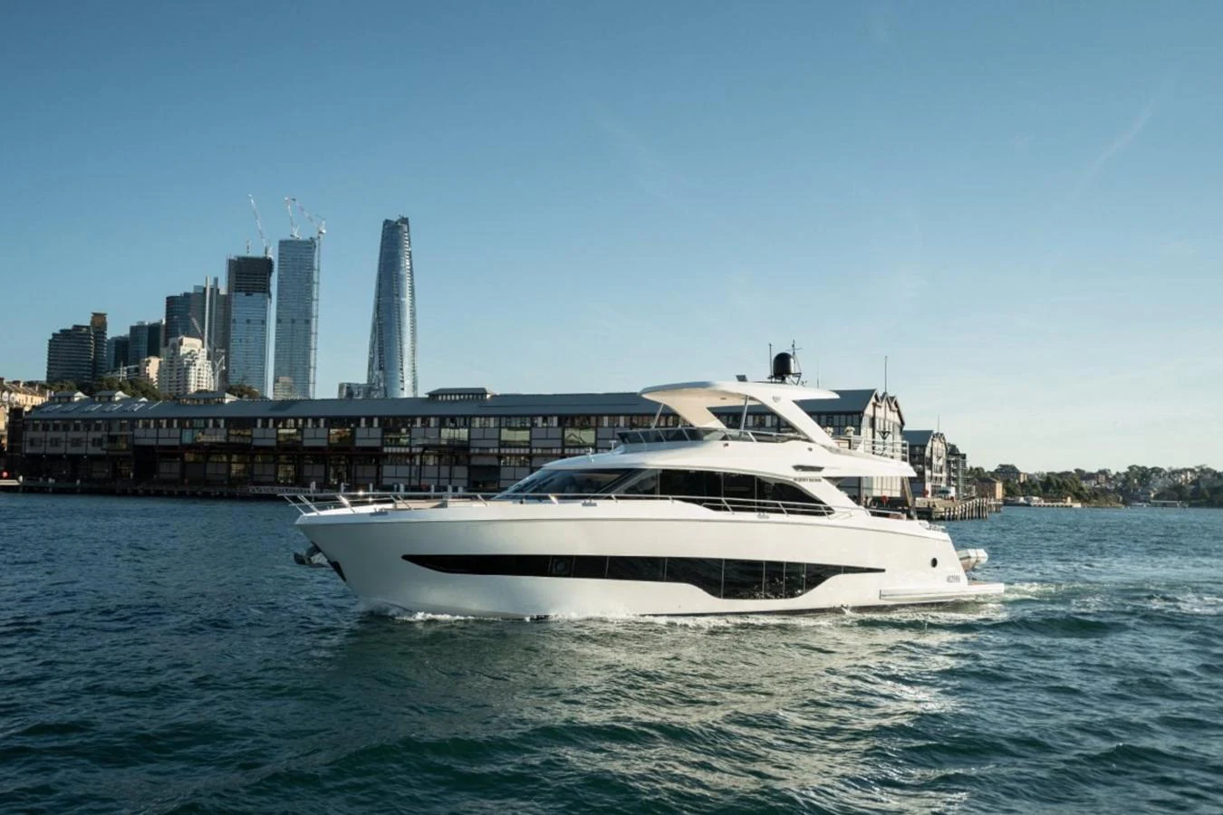 A sleek, modern yacht cruises along the waterfront with a cityscape in the background. aboard TOPAZ Yacht for Sale