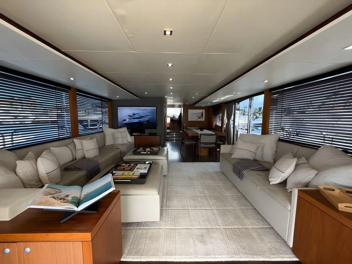 The salon features plush seating, a coffee table with books, and large windows with blinds, creating a cozy and inviting atmosphere. aboard EBRA Yacht for Sale