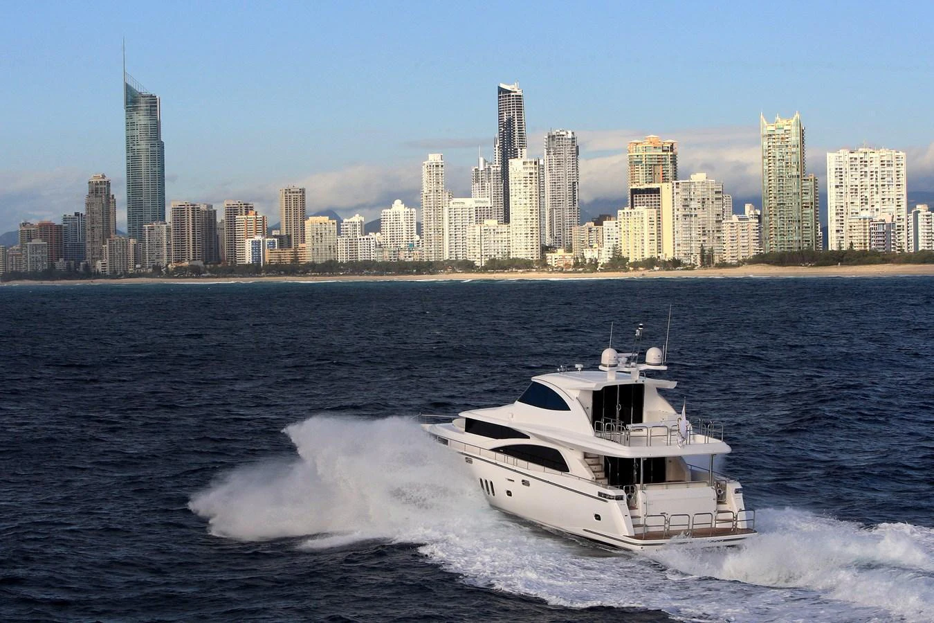 A sleek yacht cruises through the water with a city skyline in the background. aboard JOHNSON 83’ SKYLOUNGE W/FISHING COCKPIT Yacht for Sale