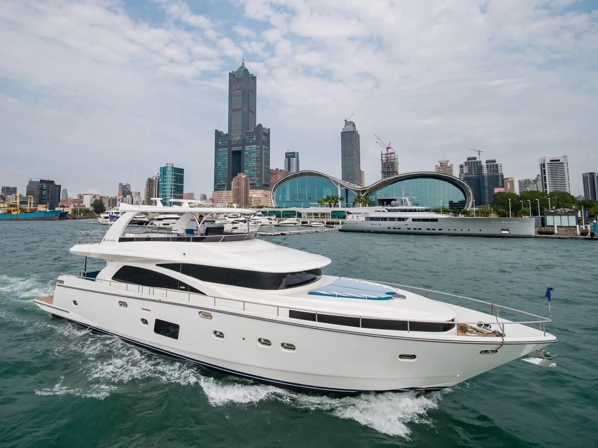 A sleek white yacht cruises through a bustling harbor with a city skyline in the background. aboard JOHNSON 83' FLYBRIDGE W/HYDRAULIC PLATFORM Yacht for Sale