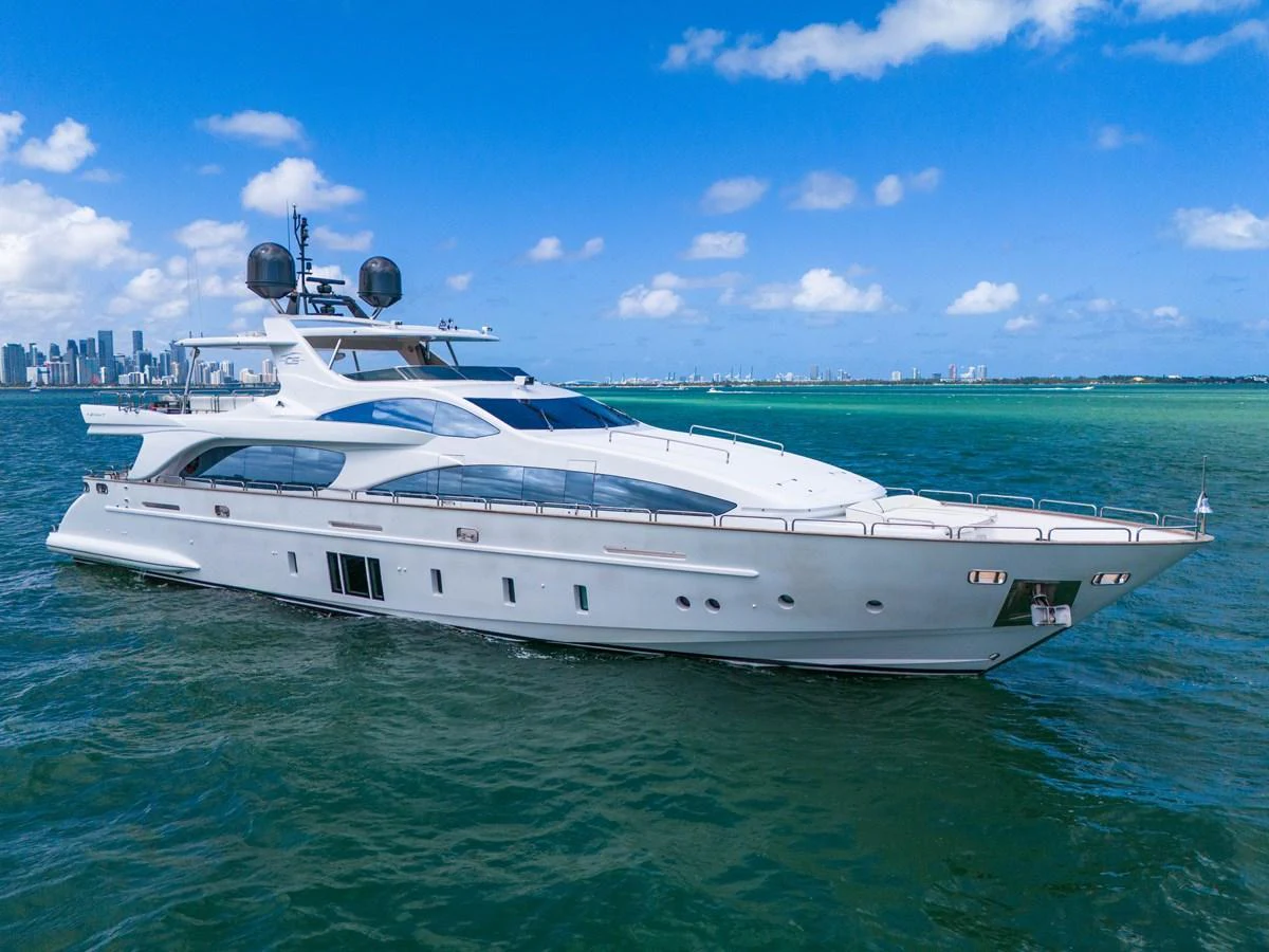A sleek and modern yacht cruising on clear blue waters with a cityscape in the background. aboard AZIMUT GRANDE 105 Yacht for Sale
