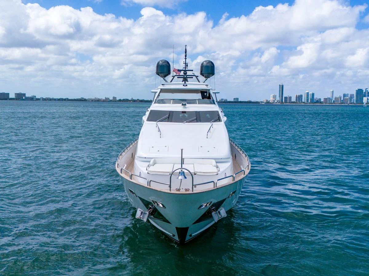 A sleek yacht is seen head-on, cruising on calm waters with a city skyline in the background. aboard AZIMUT GRANDE 105 Yacht for Sale