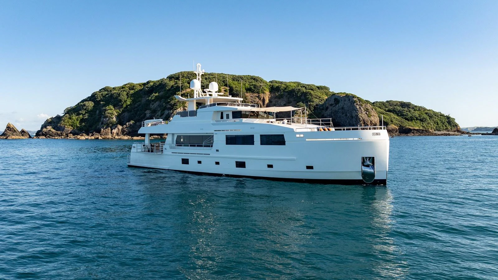 A sleek, modern yacht is anchored in calm waters with a scenic island backdrop. aboard NYLA Yacht for Sale