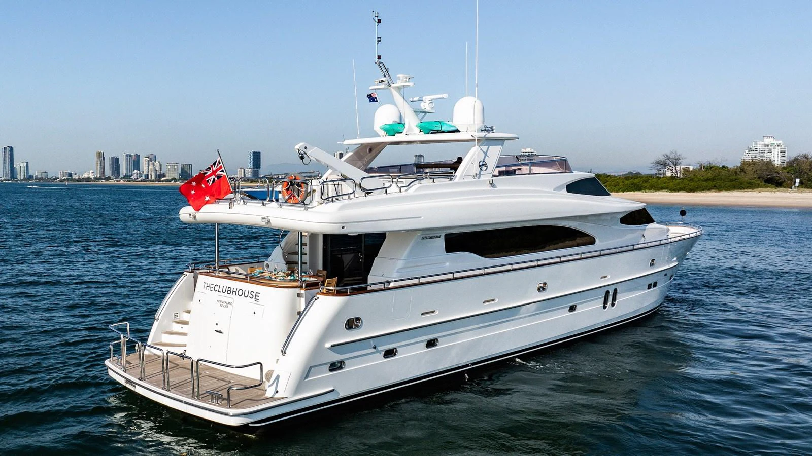 A sleek and modern yacht cruising near the coastline with a cityscape in the background. aboard THE CLUBHOUSE Yacht for Sale