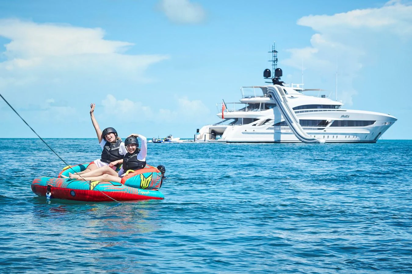 A luxurious yacht with a waterslide is anchored in a serene blue sea, while two people enjoy a fun ride on an inflatable water toy. aboard ARKADIA Yacht for Sale