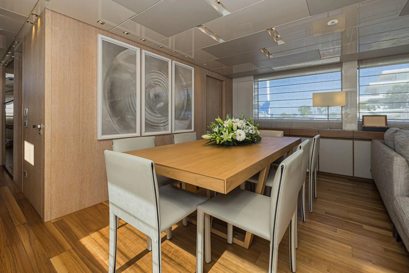 The yacht's dining area features a sleek wooden table surrounded by modern chairs, complemented by contemporary artwork and ample natural light. aboard PERCAL Yacht for Sale