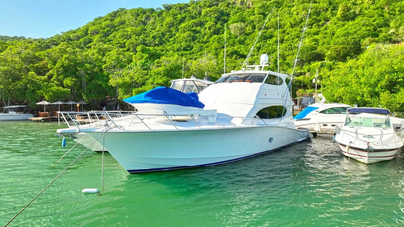 A sleek white yacht is moored in a serene, green-water bay surrounded by lush greenery. aboard HATTERAS 68 CONVERTIBLE Yacht for Sale