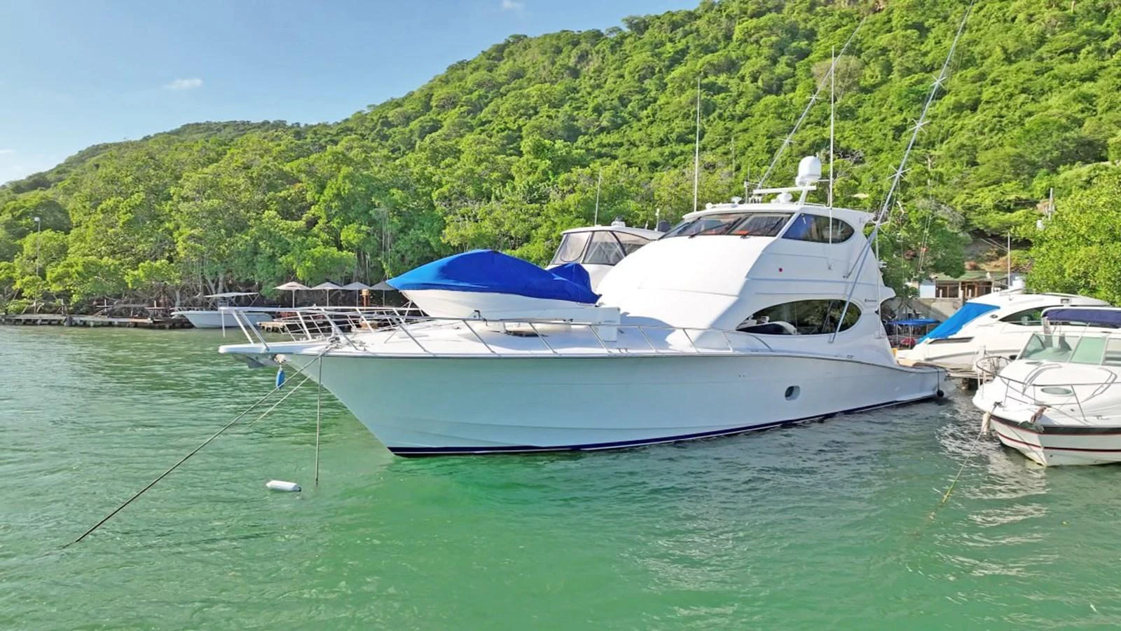 A sleek white yacht is anchored in a serene, green-water bay with lush hills in the background. aboard HATTERAS 68 CONVERTIBLE Yacht for Sale