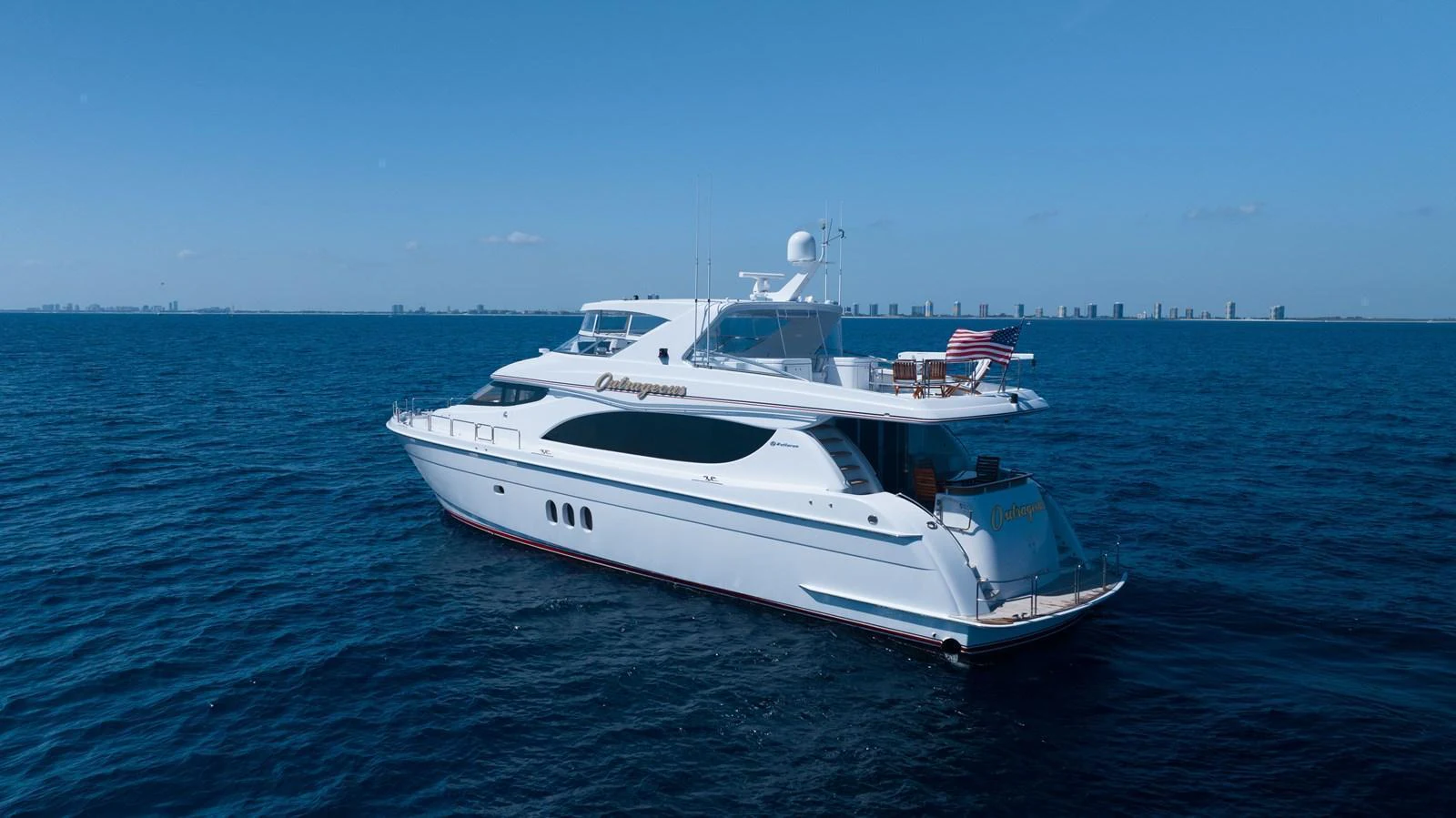 A sleek and elegant yacht cruising on calm blue waters with a distant city skyline. aboard OUTRAGEOUS Yacht for Sale