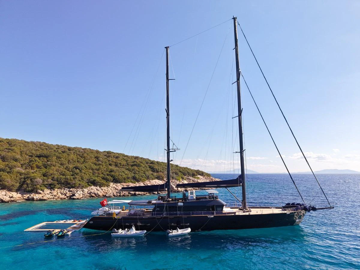 A sleek sailing yacht anchored in a serene, turquoise bay with lush greenery in the background. aboard OGUZ KHAN Yacht for Sale