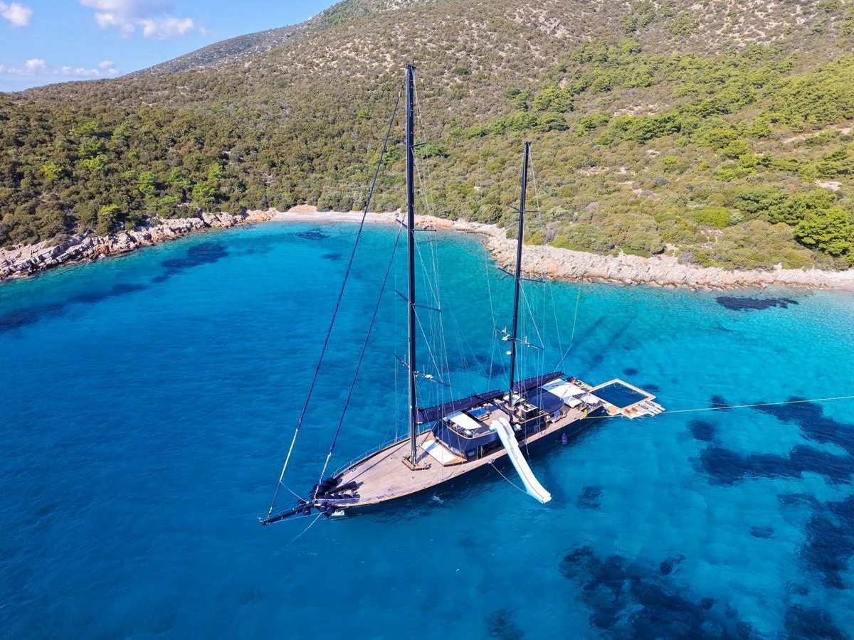 A stunning yacht anchored in a serene, turquoise bay with lush greenery in the background. aboard OGUZ KHAN Yacht for Sale