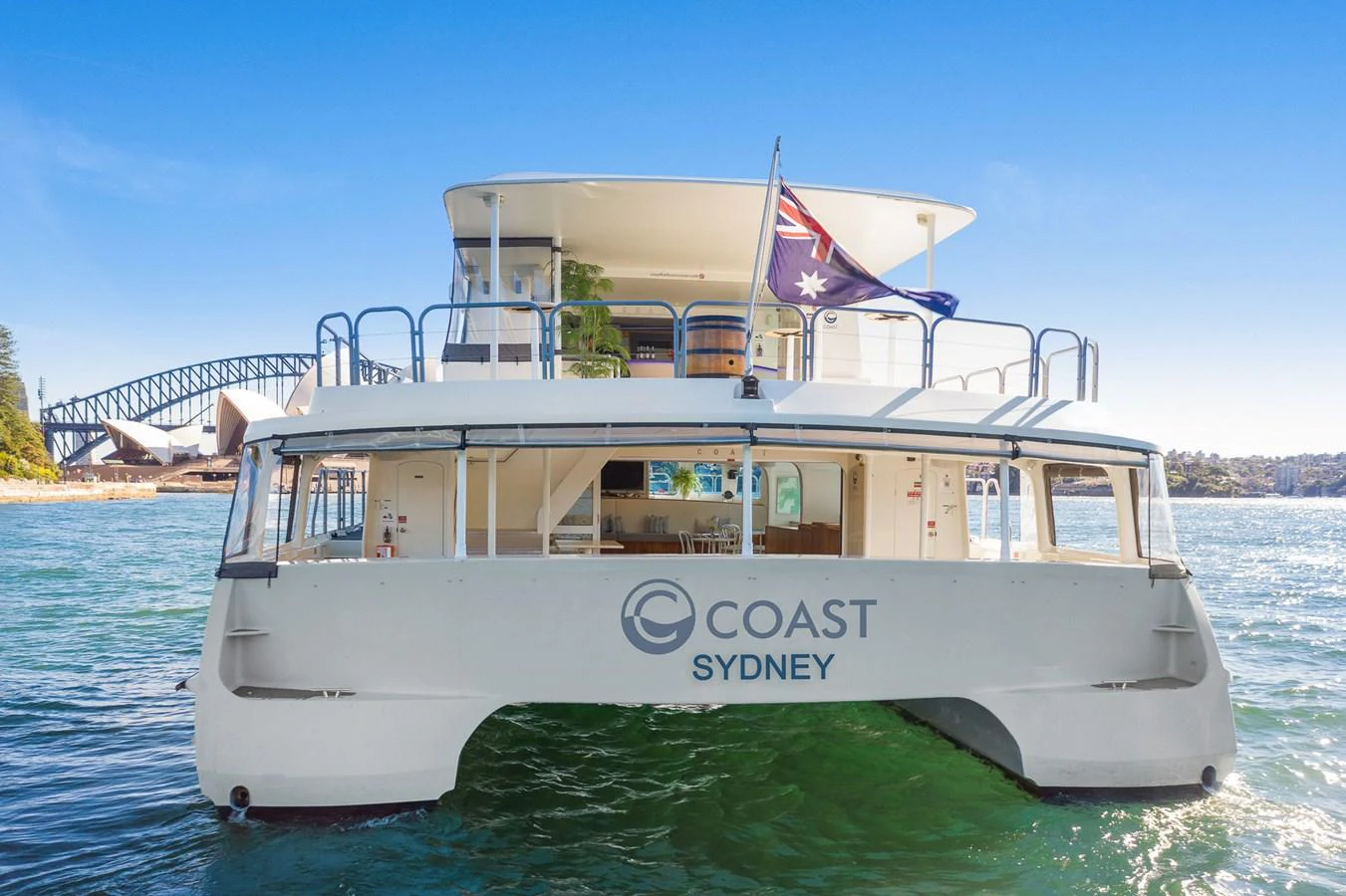 A modern catamaran yacht with spacious decks and an Australian flag, set against a scenic harbor backdrop. aboard COAST Yacht for Sale