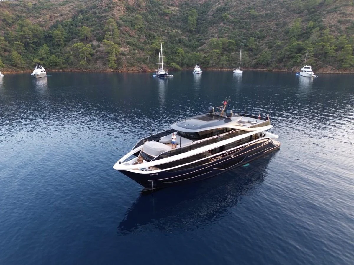 A sleek yacht cruises gracefully on calm waters with a scenic backdrop of lush hills and anchored boats. aboard OCEAN THREE Yacht for Sale