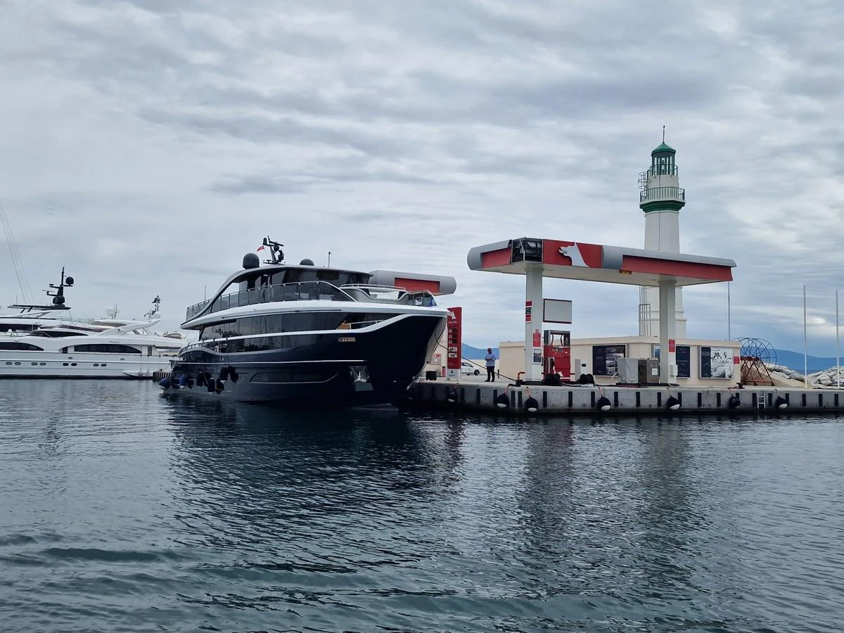 A sleek yacht is docked at a marina next to a fuel station and lighthouse. aboard OCEAN THREE Yacht for Sale