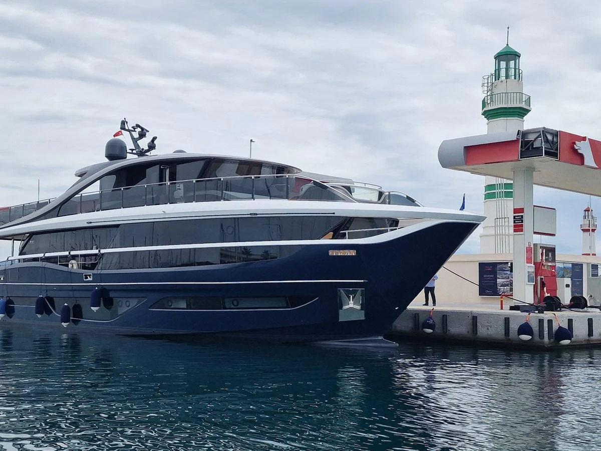 A sleek yacht is docked at a marina with a lighthouse in the background. aboard OCEAN THREE Yacht for Sale
