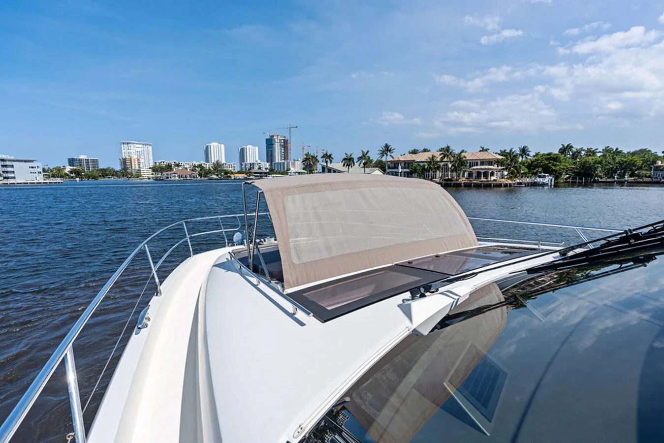 The yacht's exterior features a sleek design with a sunshade on the bow, set against a backdrop of waterfront properties and a clear blue sky. aboard JOVE Yacht for Sale
