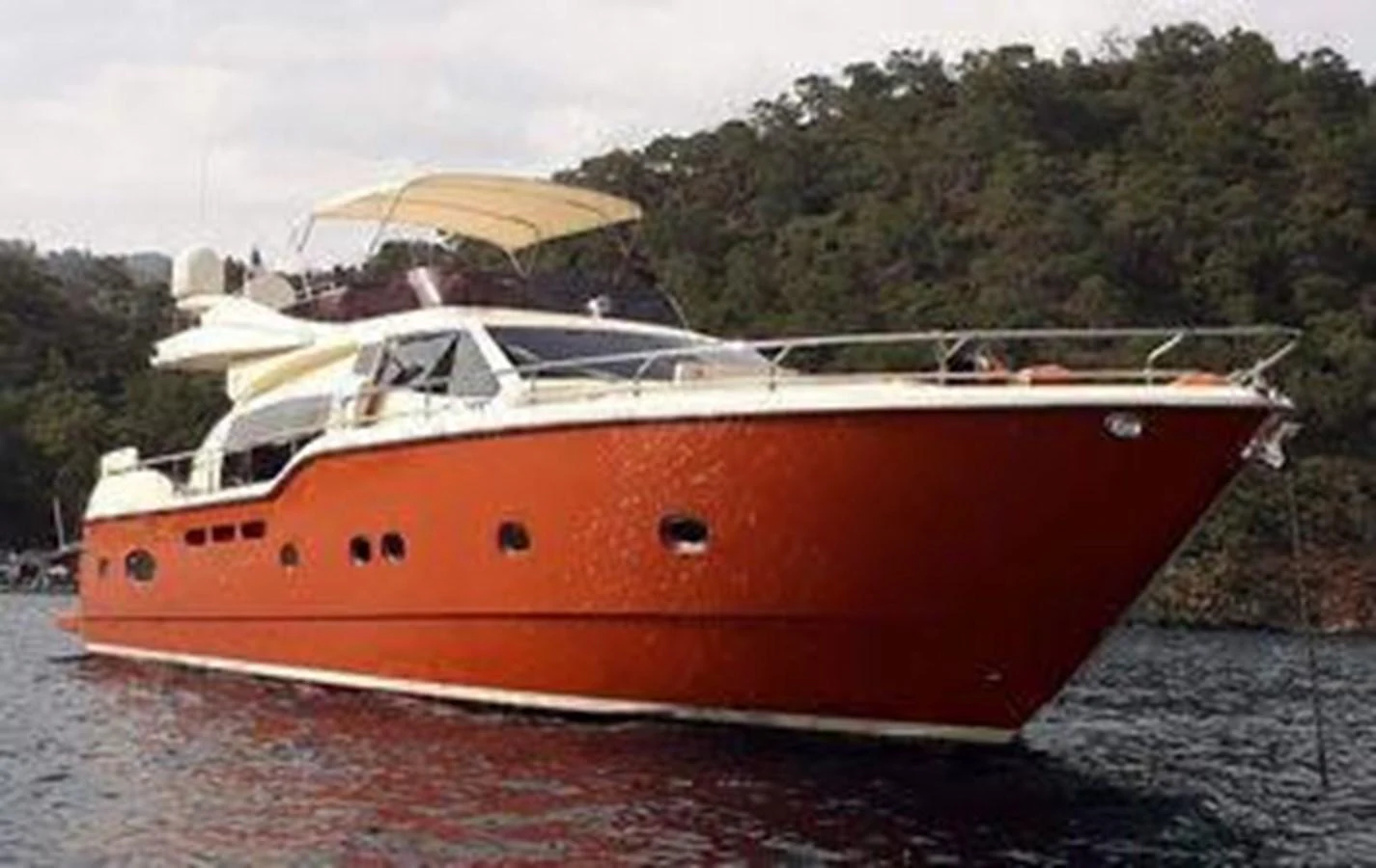 A sleek red-hulled yacht is anchored in calm waters against a backdrop of lush greenery. aboard CARAMEL Yacht for Sale