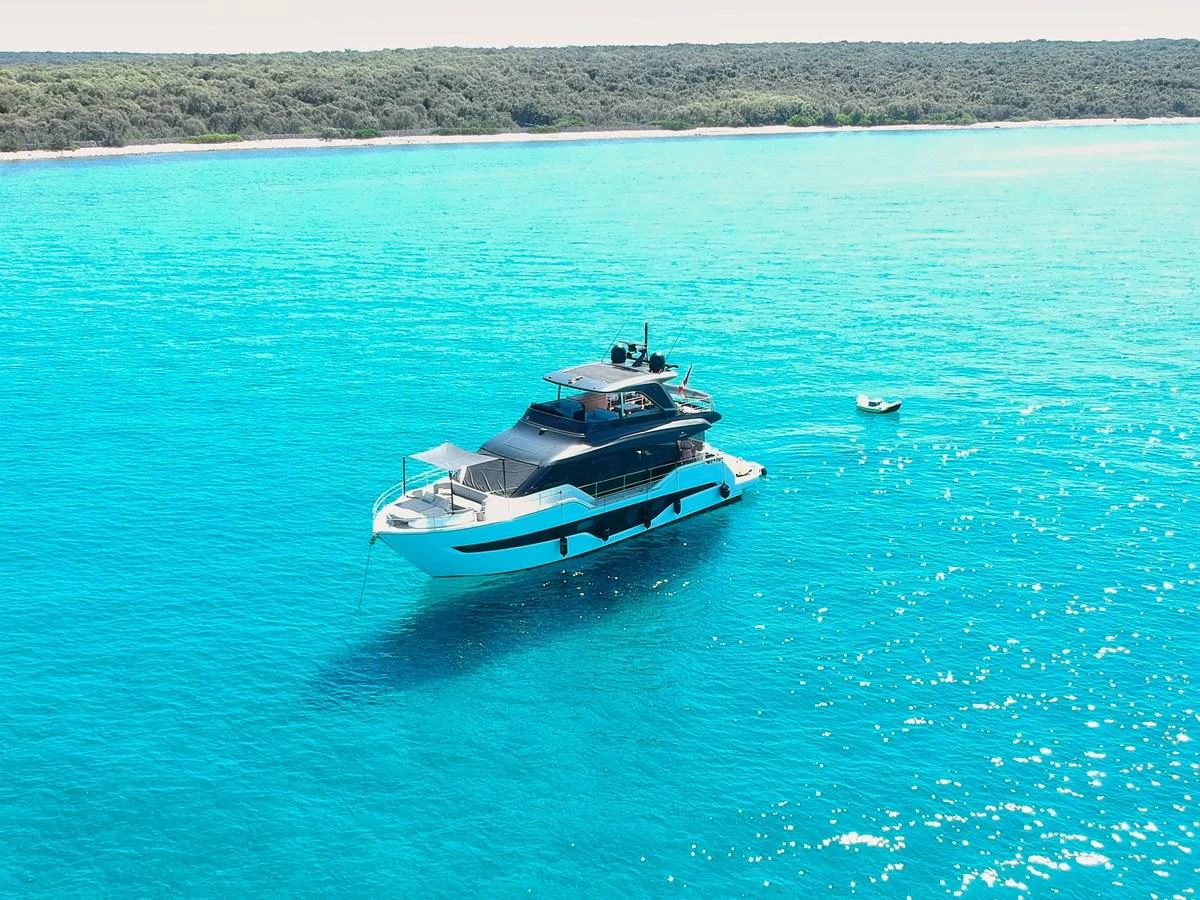 A sleek yacht is anchored in crystal-clear turquoise waters near a lush, green shoreline. aboard AMY Yacht for Sale