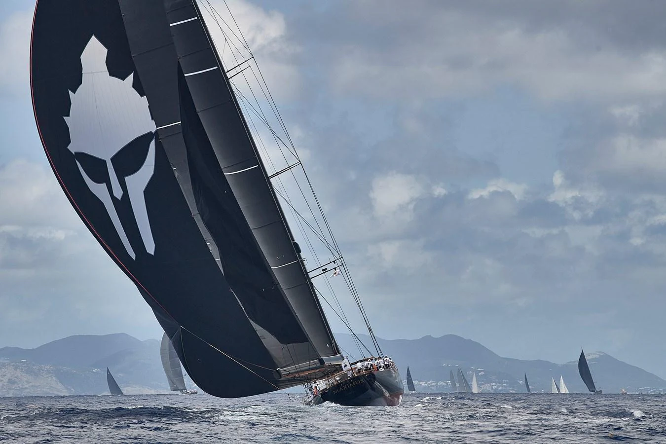 A sleek sailing yacht glides through the water with its sails fully extended, showcasing a striking design against a backdrop of distant mountains and other sailboats. aboard MAXIMUS Yacht for Sale