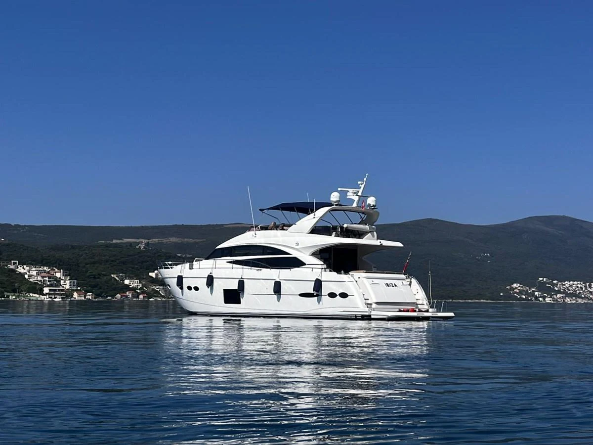 A sleek white yacht is anchored on calm waters with a scenic coastal backdrop. aboard KAZZA Yacht for Sale