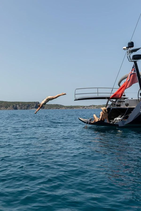 A person dives off the stern of a yacht into clear blue waters, with another person relaxing on the swim platform. aboard CAIAROSSA Yacht for Sale