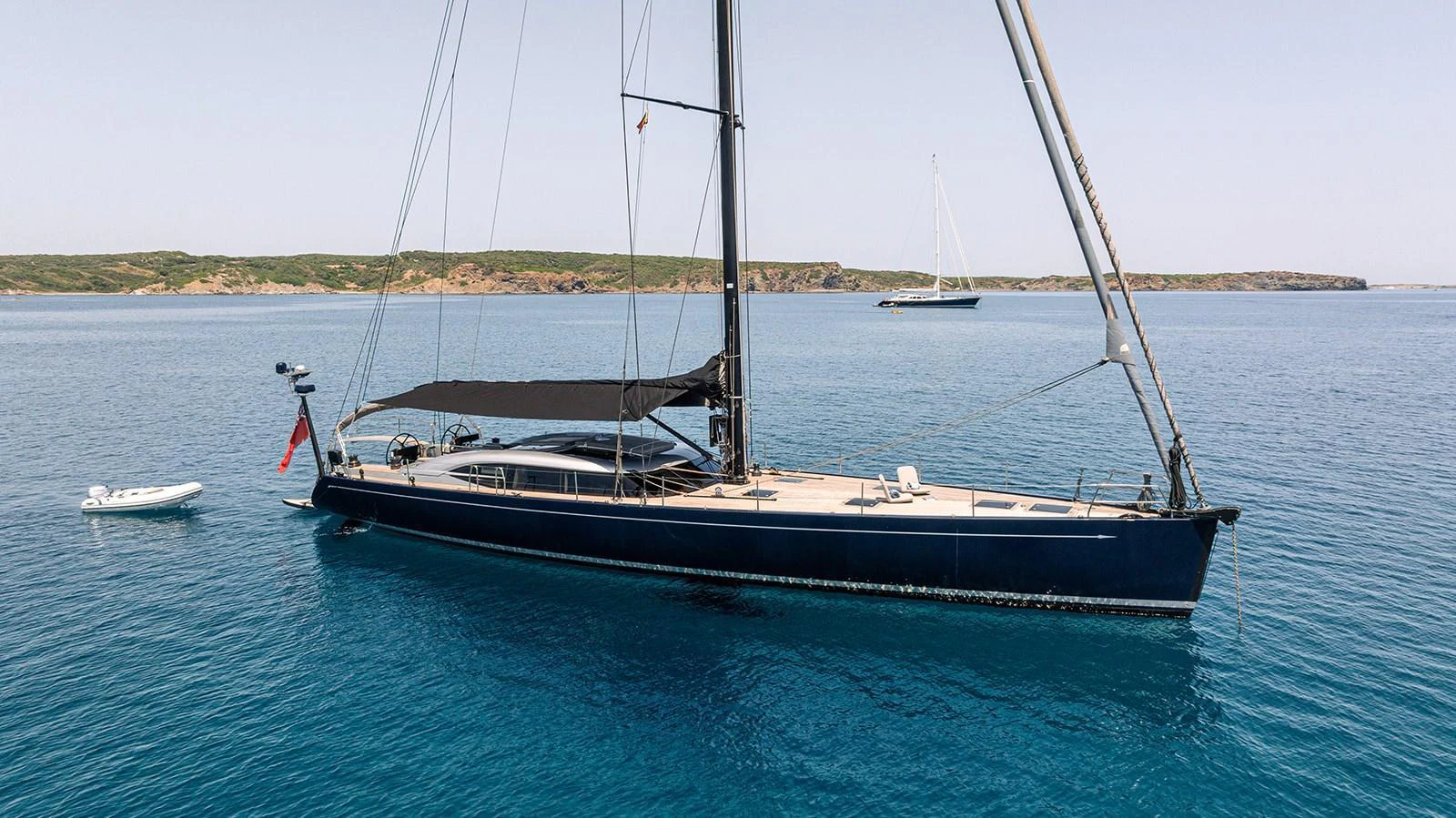 A sleek and modern sailing yacht is anchored in calm, clear blue waters with a tender nearby. aboard CAIAROSSA Yacht for Sale