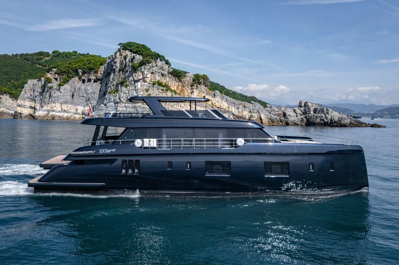 A sleek, modern yacht cruises along a scenic coastline with rocky cliffs and lush greenery. aboard PETITE BELLE Yacht for Sale