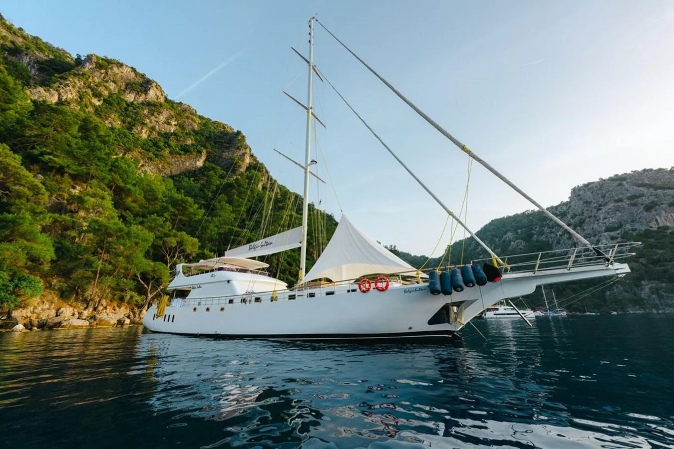 A sleek white yacht is anchored in a serene bay surrounded by lush green hills. aboard BELGIN SULTAN Yacht for Sale