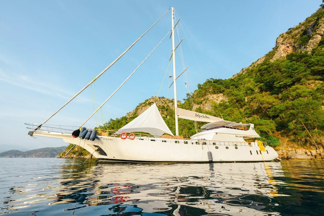 A sleek yacht is anchored near a scenic, tree-covered coastline, showcasing its elegant design and spacious deck. aboard BELGIN SULTAN Yacht for Sale
