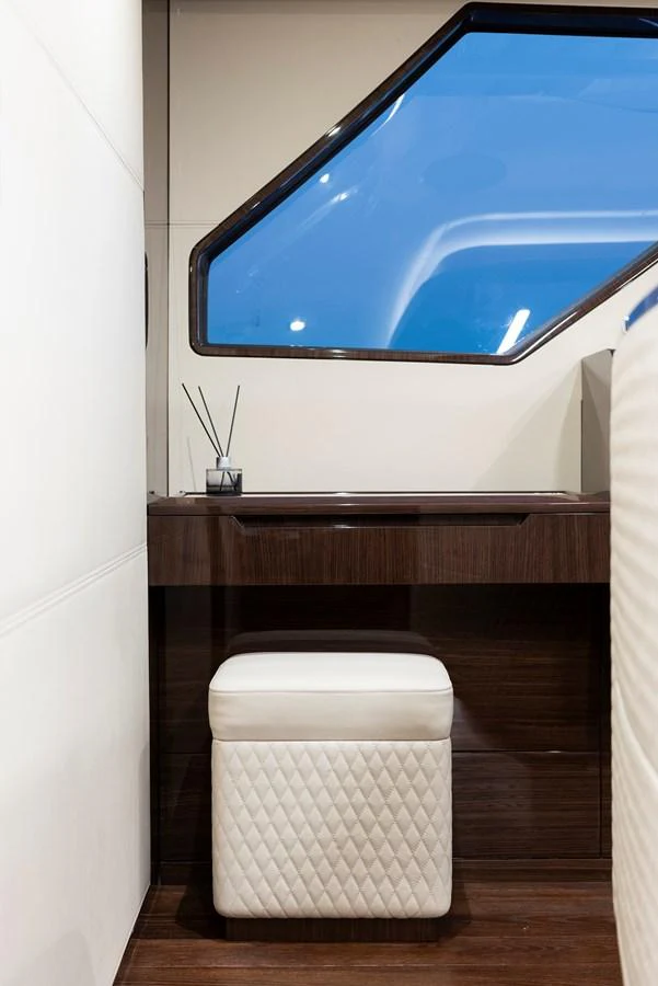 A stylish interior space featuring a sleek wooden desk and a quilted white stool, illuminated by natural light through a large window. aboard NEW BUILD Yacht for Sale