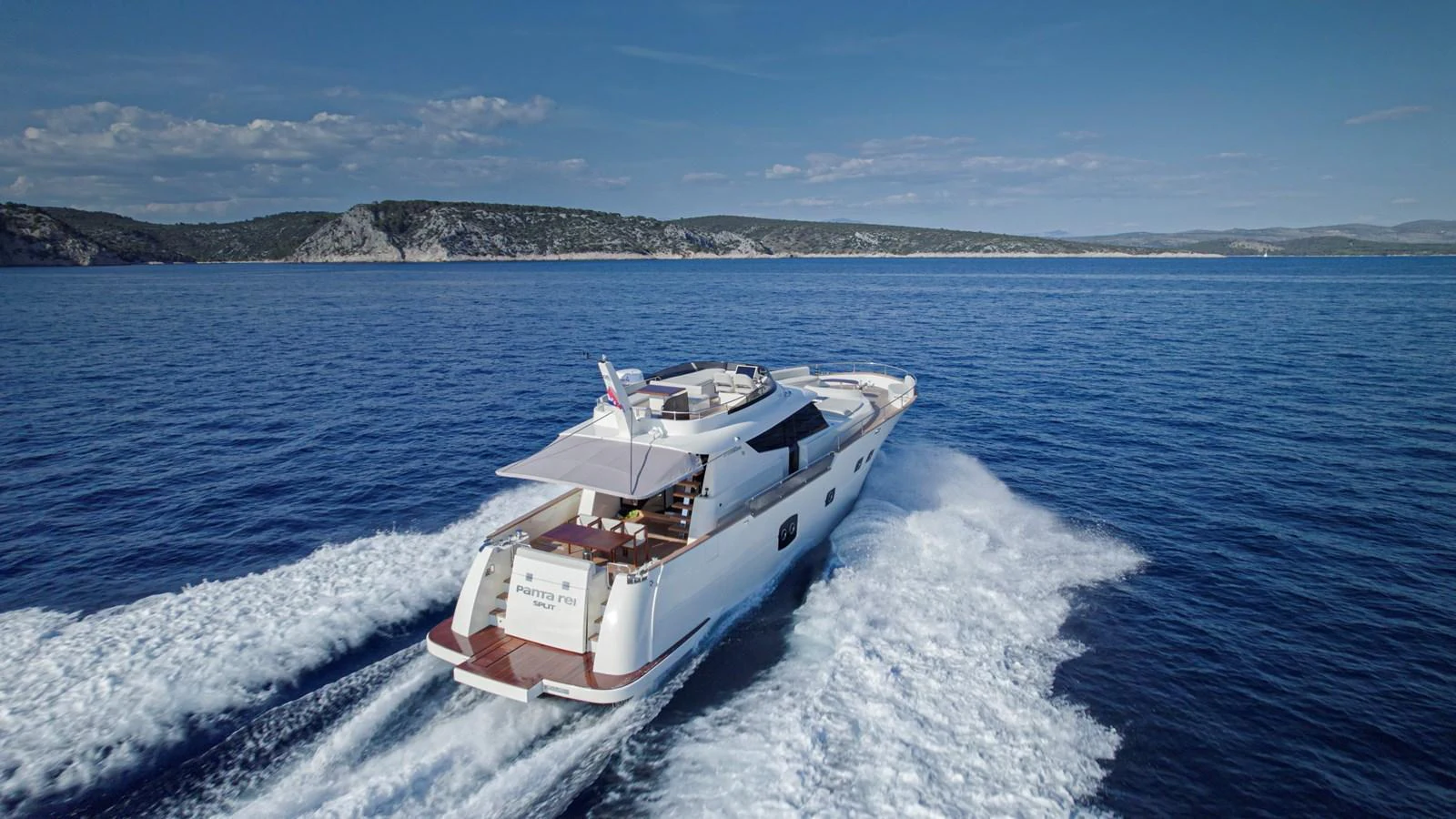 A sleek yacht cruises through clear blue waters with a scenic coastline in the background. aboard MONACHUS 70 FLY Yacht for Sale
