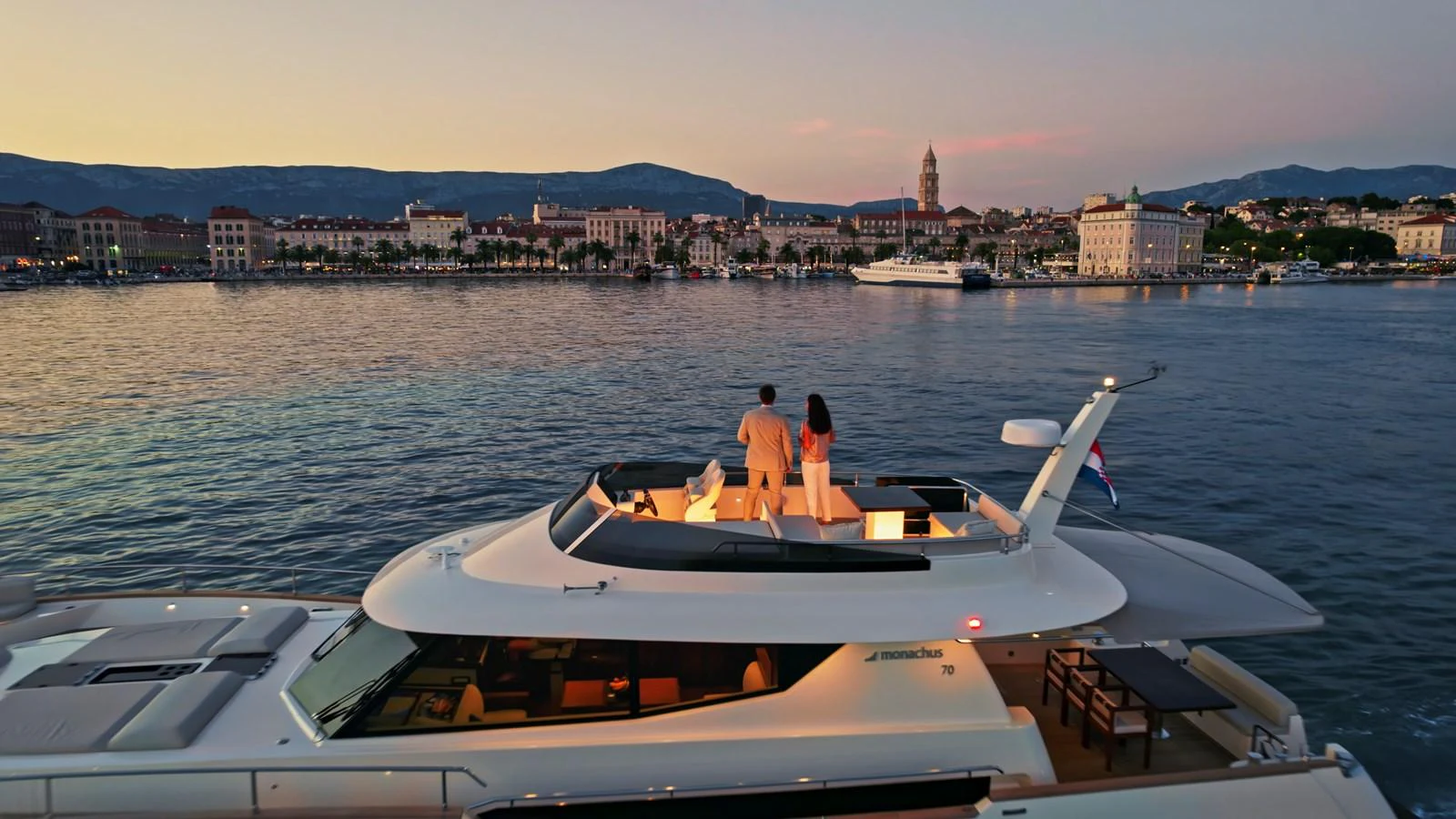 A luxurious yacht with a flybridge cruising near a picturesque coastal town at sunset. aboard MONACHUS 70 FLY Yacht for Sale