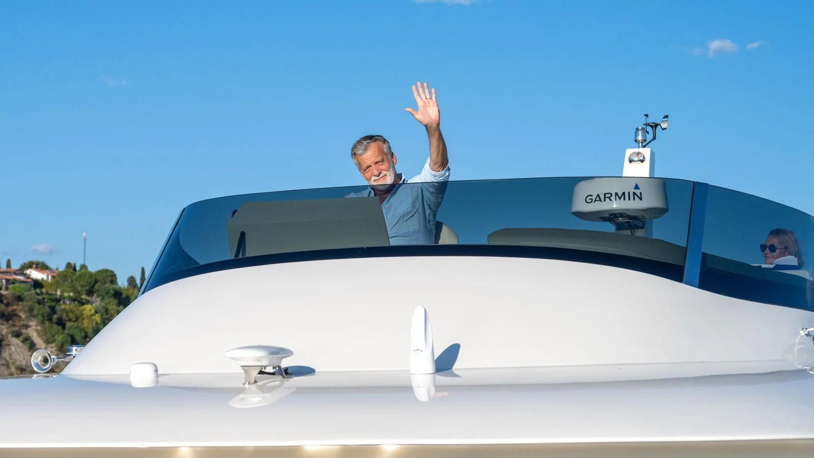 A man waves from the helm of a sleek yacht under a clear blue sky. aboard MONACHUS 70 FLY Yacht for Sale