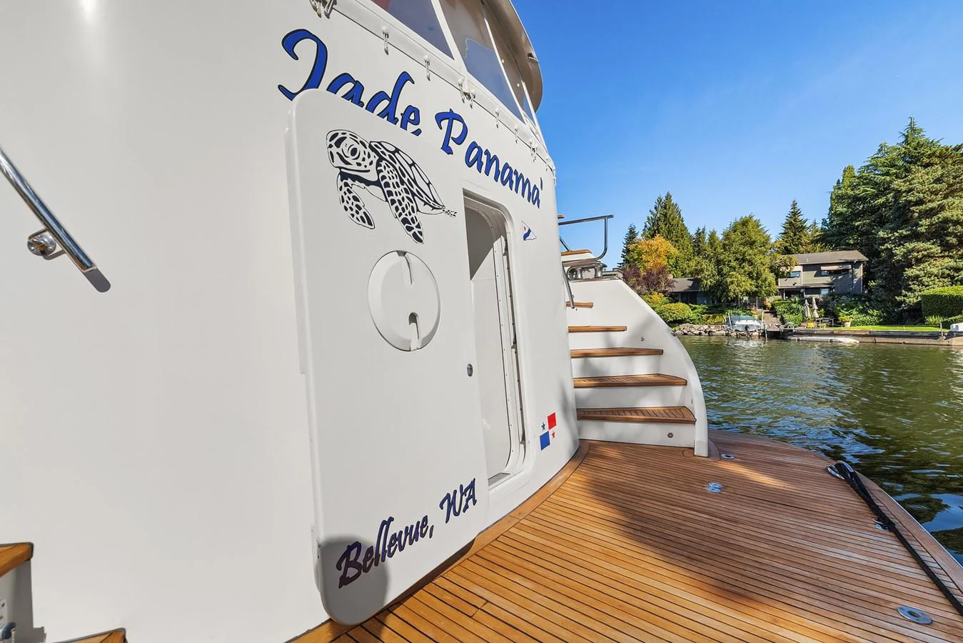 The yacht's stern features a wooden deck and a door with a turtle graphic, docked in a serene waterside setting. aboard MI AMERE Yacht for Sale