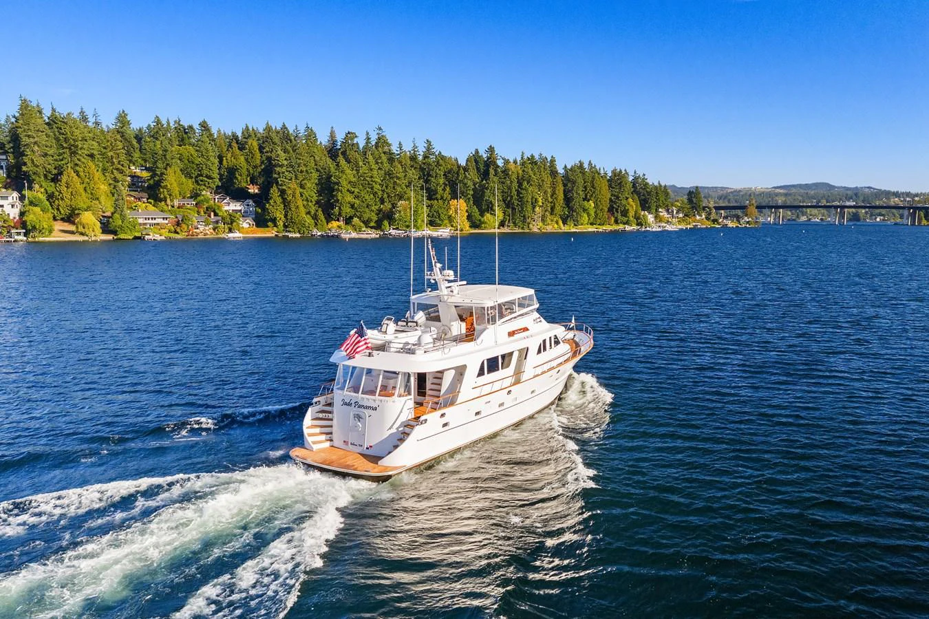 A luxurious yacht cruises through calm waters with a scenic backdrop of trees and a bridge. aboard MI AMERE Yacht for Sale