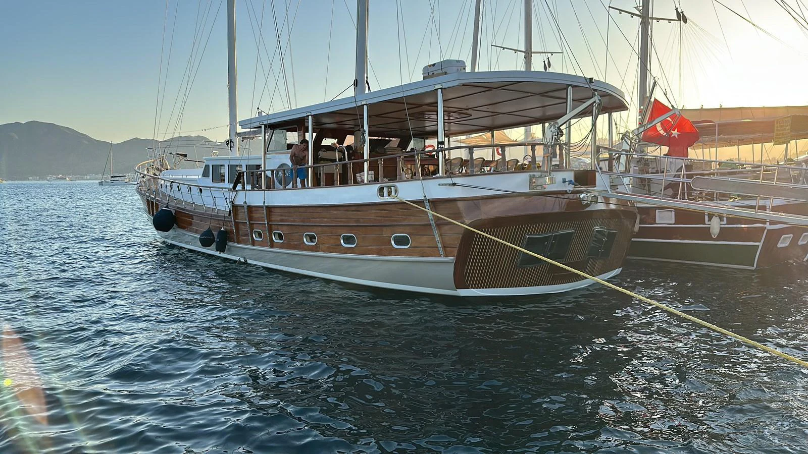 A classic wooden yacht with a spacious deck, moored in a serene harbor at sunset. aboard HANGOVER Yacht for Sale