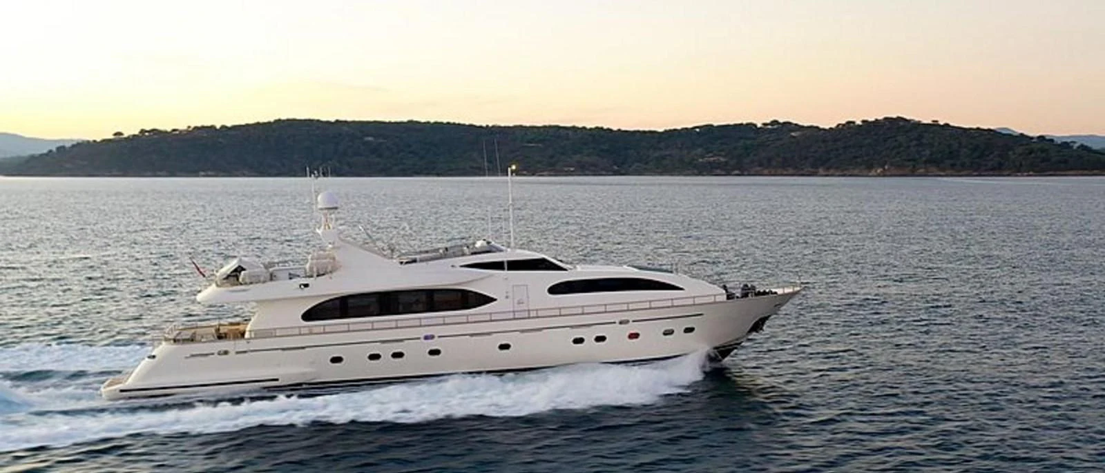 A sleek yacht glides through calm waters against a backdrop of distant hills. aboard BALKAN Yacht for Sale