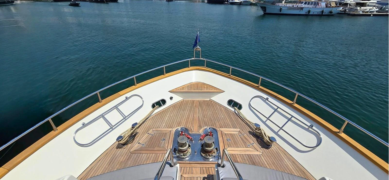 The bow of the yacht showcases a spacious deck with polished wood and stainless steel fittings, set against a serene marina backdrop. aboard BALKAN Yacht for Sale