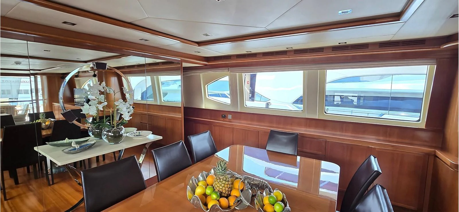 The yacht's dining area features a polished wooden table with a centerpiece of fresh fruit, surrounded by comfortable seating and large windows offering a view of the sea. aboard BALKAN Yacht for Sale