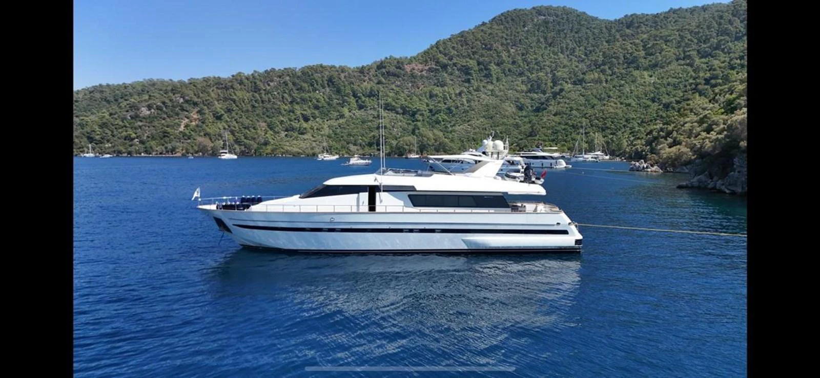 A sleek yacht cruises through calm blue waters with a lush, green hillside in the background. aboard TWINS PC Yacht for Sale