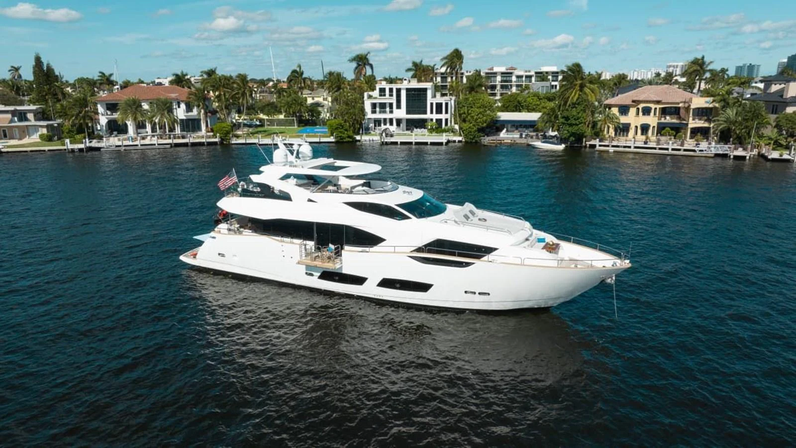 A sleek, modern yacht cruises along a serene waterfront lined with luxurious homes. aboard 95 FT 2018 SUNSEEKER 95 YACHT, MIRRACLE Yacht for Sale